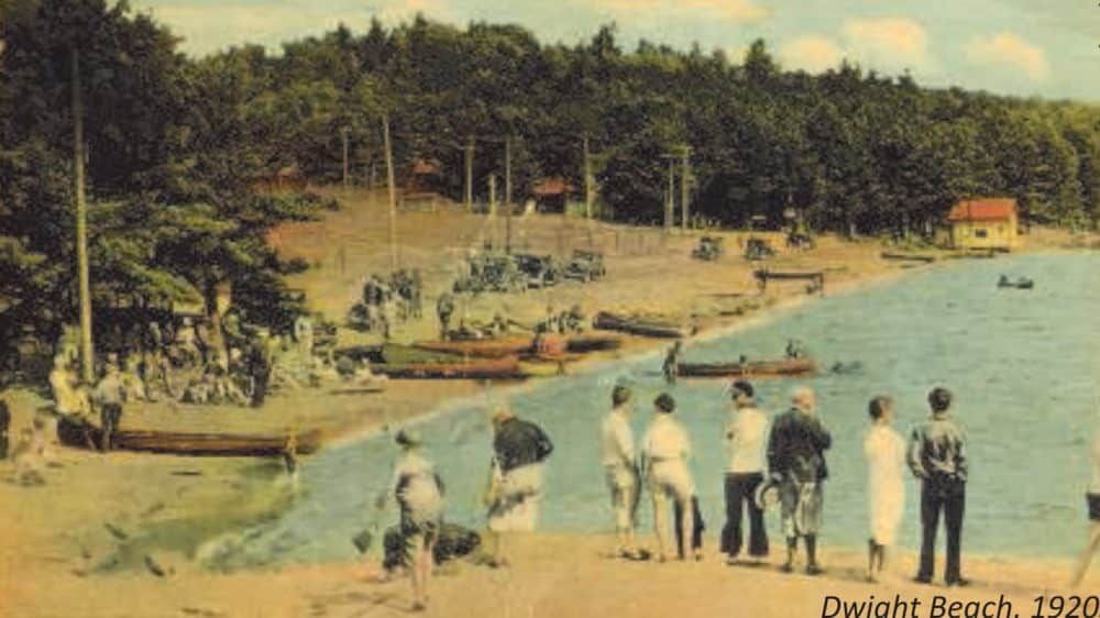 Colourized picture of a beach in 1920 with lots of people standing around and enjoying the waterfront in boats in Muskoka